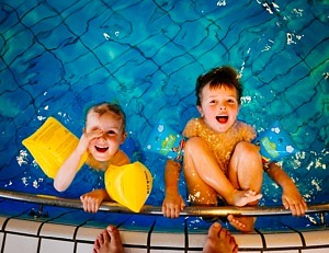 Två barn i en bassäng. Håller i kanten och ler in i kameran.